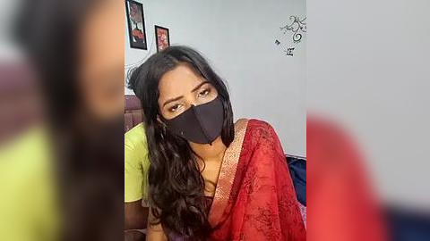 Media: A video of a woman in a red saree with a black mask, sitting in a room with framed pictures and a red wall.