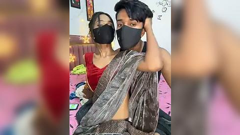 Media: A video of two young, South Asian individuals in a dimly lit room, wearing black masks, red and gray clothing, on a pink bed, blurry background.
