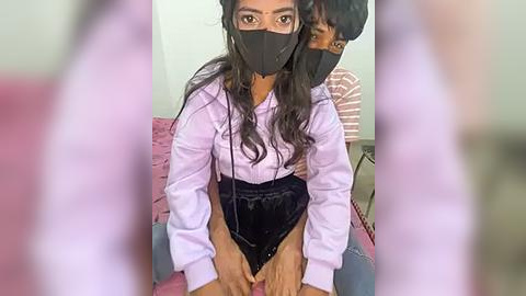 Media: Video of two women with South Asian features, wearing black face masks, light lavender tops, and high-waisted shorts. One sits on a pink couch, the other stands behind her.