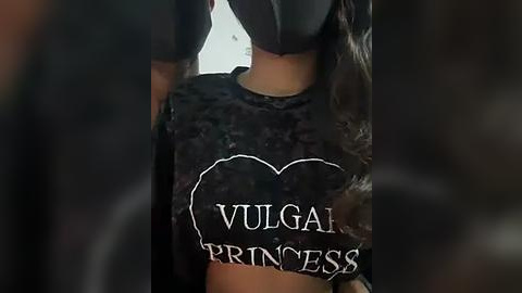 Media: Video of a person wearing a black, velvet-like shirt with the words \"VULGAR PRINCESS\" in white text, partially obscured by their hands. The background is blurred and dark, emphasizing the bold, edgy fashion statement.