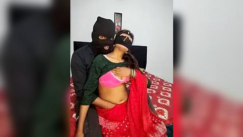 Media: Video of a woman in a red saree and green blouse, wearing a black mask, being held by a man in a black shirt, in a bedroom with a bed covered in a patterned red blanket.