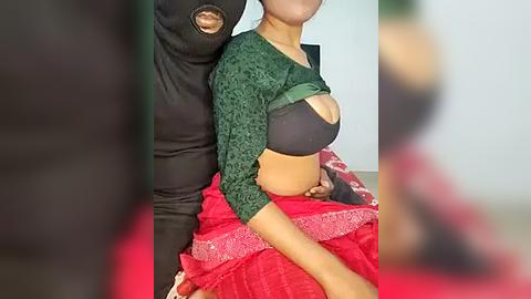 Media: Video of a woman with a dark complexion, wearing a green cardigan and black bra, sitting on a red blanket, with a man's arm around her waist.