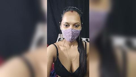 Media: Video of a woman with medium brown skin, wearing a purple face mask, black bra, and long dark hair, seated in a black chair, blurred background.