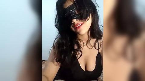 Media: Video of a smiling woman with long black hair, wearing a black lace mask and black bra. Her skin is light, and she has a curvy physique. The background is a plain light blue wall.