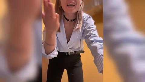 Media: Video of a woman in a light blue button-up shirt and black pants, with a black choker, smiling and waving with both hands, set against a blurred background.