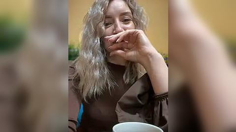 Media: Video of a middle-aged woman with long, wavy gray hair, wearing a brown top, eating a sandwich with her right hand, blurred background, yellow wall, and a white cup in the foreground.