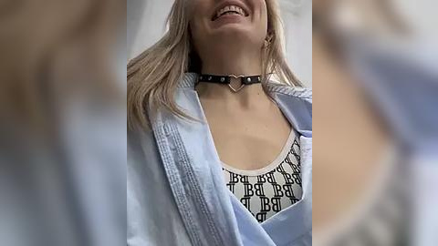 Media: Video of a smiling, fair-skinned, blonde woman wearing a black studded choker, a white shirt with black \"BRA\" pattern, and a light blue denim jacket.