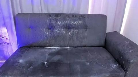 Media: Video of a dark gray, leather couch with a tufted backrest, heavily soiled with dirt and stains, positioned against white curtains in a dimly lit room.