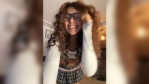 Media: Video of a smiling, curly-haired woman in glasses, wearing a white top and plaid skirt, in a warmly lit room with framed art and a hanging lamp.