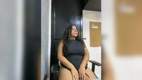 Media: A video shows a plus-sized Latina woman with curly hair in a black bodysuit, sitting in a dimly lit room with a black door and a whiteboard in the background.