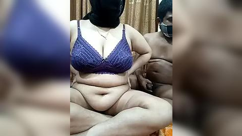 Media: Video of a plus-sized, light-skinned woman wearing a blue lace bra and black hijab, seated on a bed with a dark-skinned man partially visible behind her.