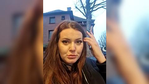 Media: Video of a Caucasian woman with long, straight brown hair, wearing a black jacket, with a puzzled expression, taken outdoors near a brick building and bare tree.