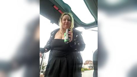 Media: Video of a blonde woman in a black dress and coat holding a can inside a car, blurred people in the foreground, suburban background.