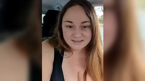 Media: A video of a young, light-skinned woman with long brown hair, smiling slightly, wearing a black tank top, seated in a car. The background is blurred.