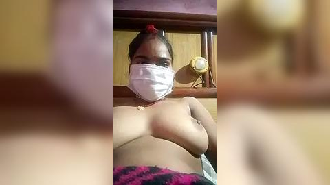 Media: Video of a topless, light-skinned woman with medium breasts, wearing a red headband, white mask, and pink and black plaid blanket. Background includes wooden shelves and a yellow alarm clock.