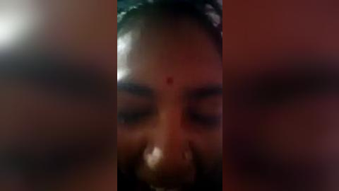 Media: A close-up video of a woman's face with a prominent red dot on her forehead, her eyes closed, and her mouth open, showing teeth. The image is blurred, making details difficult to discern.