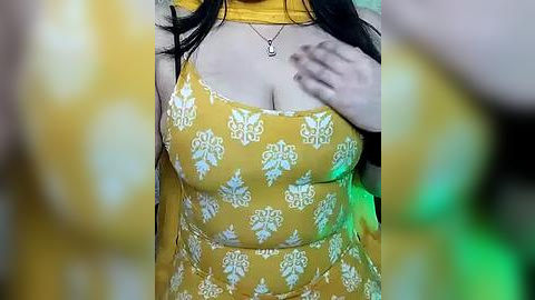 Media: Video of a woman with medium skin tone, wearing a mustard-yellow dress adorned with white floral patterns, her breasts partially visible under the dress, her long black hair partially visible, and her hand near her chest.