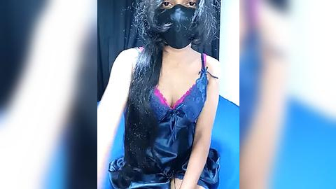 Media: Video of a young woman with long, black hair, wearing a black mask, blue satin lingerie, and sitting on a blue cushion. Background is dark with a blurred person in the foreground.