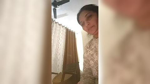 Media: Video of an Indian woman with medium brown skin and dark hair, wearing a white lace dress, standing in a dimly lit room with beige curtains and a fan on the ceiling.