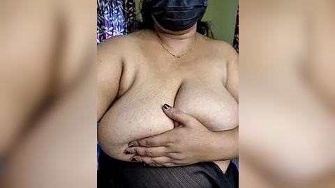 Media: A video of a topless woman with a medium build, wearing a mask and holding her large breasts with one hand. She has a gold chain necklace and dark, patterned clothing.