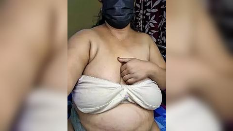 Media: Video of a plus-sized woman with medium skin tone, wearing a gray mask, white strapless bra, and blue shorts. She is covering her large breasts with her right hand. Background features a floral-patterned curtain.