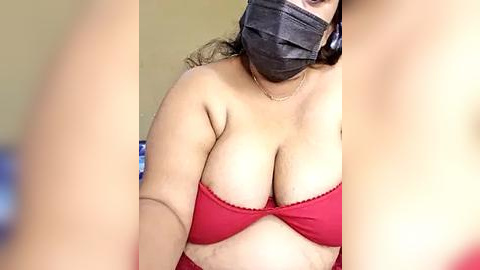 Media: Video of a plus-sized woman with a mask, wearing a red strapless bra, revealing ample cleavage, standing indoors.