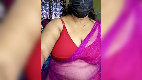 Media: Video of a woman wearing a red bra and a semi-transparent purple saree, with a black mask covering her face, in a dimly lit room.