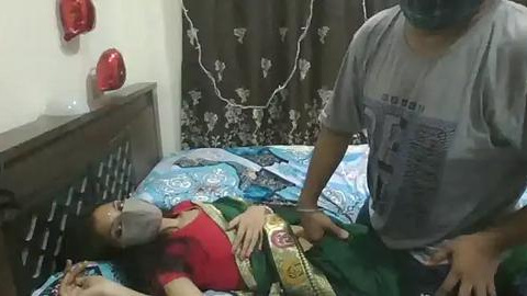 Media: Video of a woman in a gray shirt and mask attending to a child in a bed, wearing a red shirt and gray mask, in a modest bedroom with floral curtains and a wooden headboard.