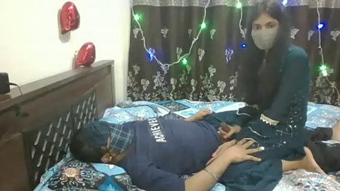 Media: Video of a woman in a mask, dark dress, and gloves comforting a man on a bed, surrounded by festive lights and curtains.