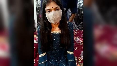 Media: Video of a young South Asian girl with long black hair, wearing a dark blue, embroidered dress, a white face mask, and a bindi on her forehead, seated on a red, floral-patterned couch.