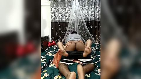 Media: Video of a dark-skinned woman lying on a floral bedspread, wearing a grey top and black shorts, with legs spread apart, under a white net canopy.