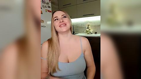 Media: A video of a smiling, fair-skinned woman with long blonde hair, wearing a light blue spaghetti-strap top, sitting in a modern kitchen with beige cabinets and a tiled backsplash.