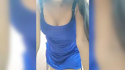 Media: Video of a woman with long, dark hair, wearing a tight, blue tank top that accentuates her large breasts, standing indoors on a carpeted floor.