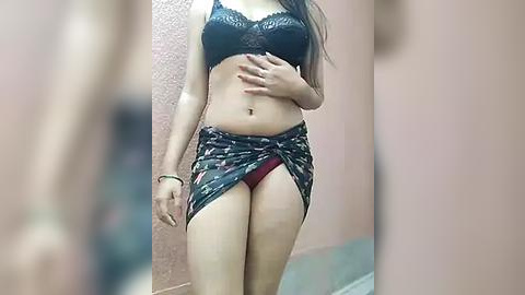 Media: Video of a slender, light-skinned woman with long dark hair, wearing a black lace bra and a colorful sarong, partially covering red panties.