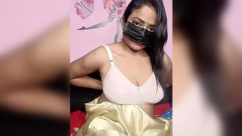 Media: Video of a South Asian woman with medium skin tone, wearing a black face mask, white bra, and beige cloth. She sits on a bed with pink walls and a heart-shaped decoration.