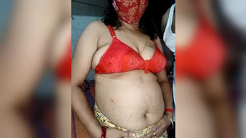 Media: Video of a pregnant South Asian woman wearing a red lace bra, red face mask, and yellow belt. Her skin is light brown, and she has a round belly. Background is blurred.