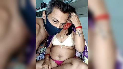 Media: Video of a shirtless man wearing a mask, adjusting a woman's mask. She wears a white bra and pink panties, sitting on a bed with a colorful patterned sheet.