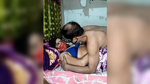 Media: Video: A shirtless, bald man in glasses kissing a woman in a blue sari on a bed with pink floral sheets in a rustic room with peeling white walls.