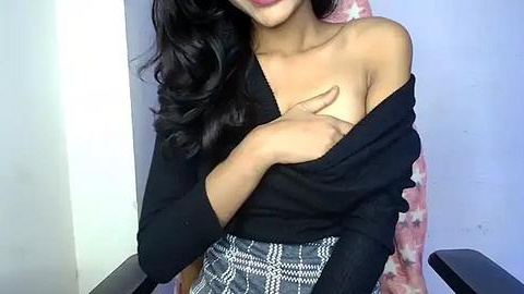 Media: Video of a woman with medium brown skin, long wavy black hair, wearing a black off-shoulder sweater and a patterned skirt, covering her chest with her hand. Background features a pink star-patterned blanket and a white wall.