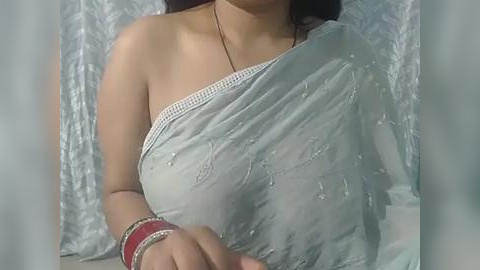 Media: A video of a South Asian woman in a light grey, embroidered saree, wearing a red bangle on her wrist, with a blurred, textured white background.