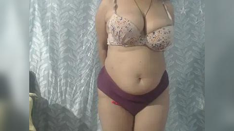 Media: Video of a plus-sized woman in a light pink floral bra and purple panties, standing in front of a silver and white patterned curtain. Her skin is light and she has a round belly.