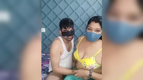 Media: Video of two people in a dimly lit room, wearing face masks and cloth face coverings, one in a yellow floral bra, sitting on a bed with a blue headboard and patterned sheets.