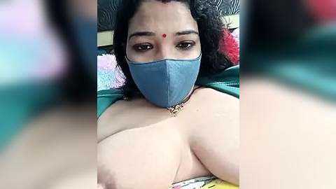 Media: Video of a South Asian woman with medium-dark skin, wearing a blue face mask and a red bindi, her curly black hair visible. She has large breasts exposed, lying on a colorful bedspread, with blurred background.