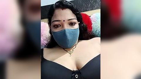 Media: Video of a South Asian woman with medium brown skin, wearing a blue surgical mask, black bra, and a gold necklace, lying on a colorful, patterned pillow, background blurred.