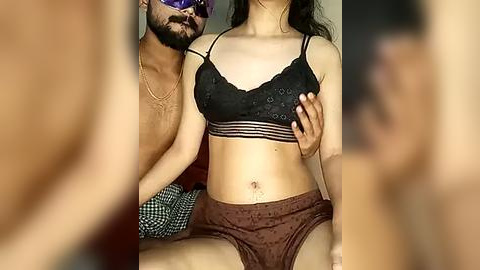 Media: Video of a topless, bearded man with a light complexion, wearing sunglasses, and a black bra, holding a slender woman with medium skin tone and long dark hair, wearing brown lace panties, seated on a couch.