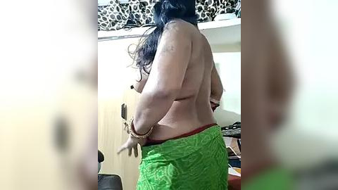 Media: Video of a topless woman with long black hair, wearing green shorts and a red thong, standing with her back to the camera, in a bedroom with a leopard-printed ceiling and wooden wardrobe.