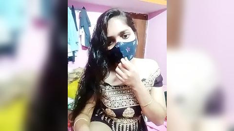 Media: Video of a young South Asian woman with long black hair, wearing a blue mask, a brown off-shoulder blouse with intricate embroidery, and a black skirt. Background shows a pink wall, clothes hanging, and a bed.