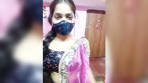 Media: Video of a young woman with medium skin tone and dark hair, wearing a black face mask, a black bra, and a pink and white saree, standing in a pink-painted room with a wooden wardrobe in the background.