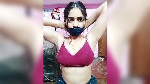 Media: Video of a young South Asian woman with medium skin tone, wearing a red sports bra and black face mask, standing in a cluttered, pink-walled room with a cartoon character poster on the wall.