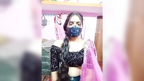 Media: Video of a South Asian woman with long, dark hair in a braid, wearing a black off-shoulder top, pink dupatta, and blue mask, seated indoors against a pink wall with hanging clothes and a wooden shelf.